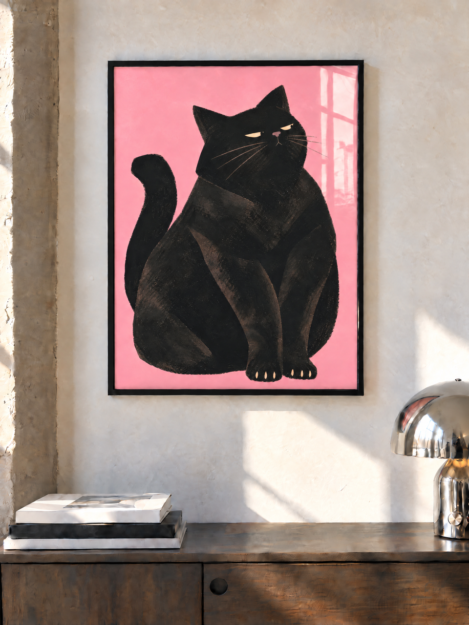 Schwarze Katze vor rosa Hintergrund in einem schwarzen Rahmen an einer sonnigen Wand über einem Holzschrank, neben Büchern und einer Chromlampe.