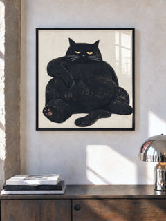 Stampa di un grosso gatto nero su fondo beige in una cornice nera, sopra una credenza in legno con libri e una lampada cromata.