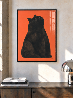 Una lámina de un gato negro sobre naranja, con marco negro, cuelga sobre un aparador de madera junto a una lámpara cromada en una habitación soleada.