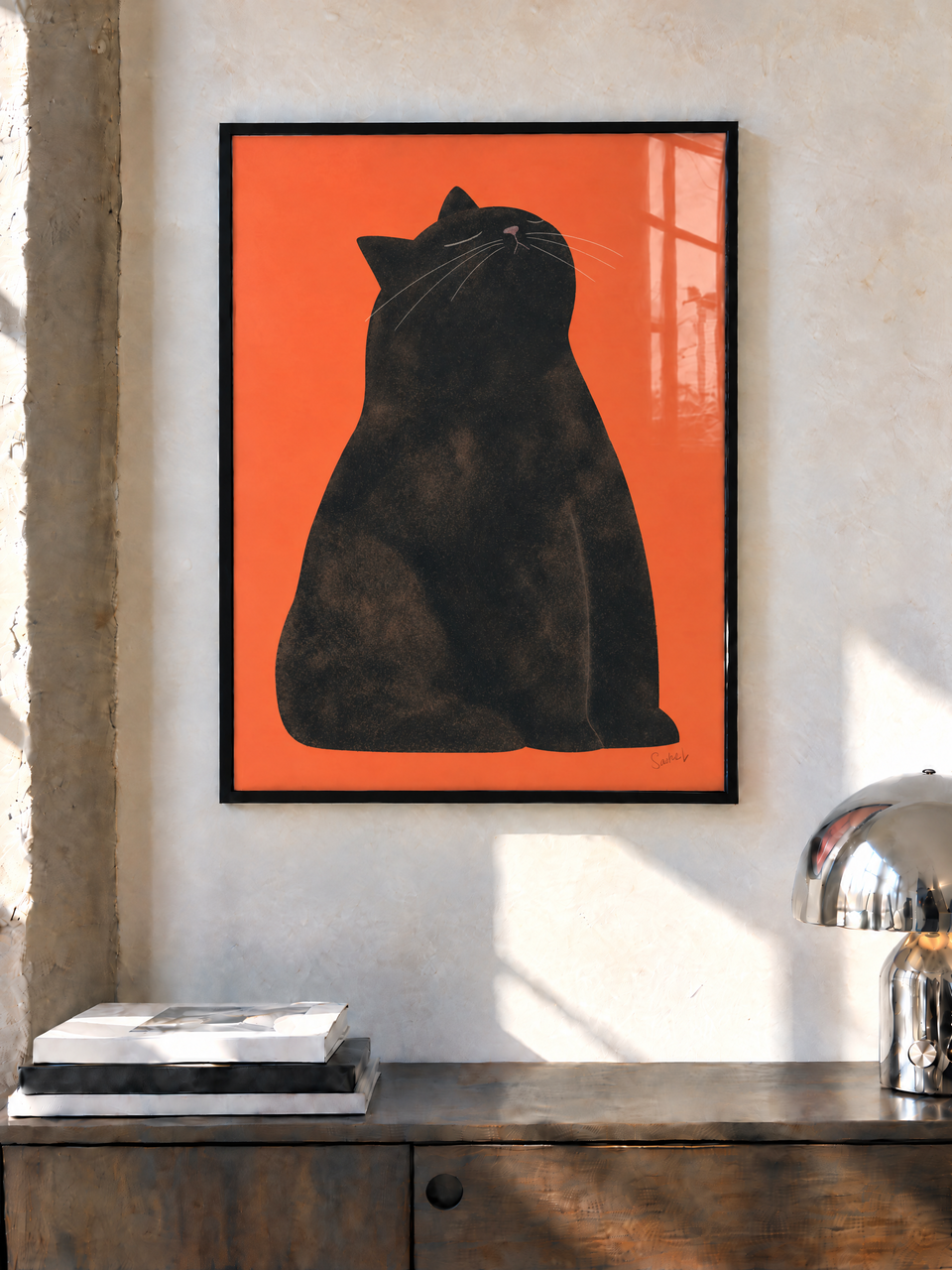 Una lámina de un gato negro sobre naranja, con marco negro, cuelga sobre un aparador de madera junto a una lámpara cromada en una habitación soleada.